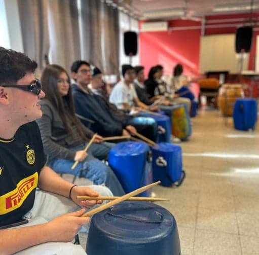 Estudiantes sentados en fila tocan con palillos tambores hechos con bidones de plástico azul, en un salón con parlantes en lo alto.